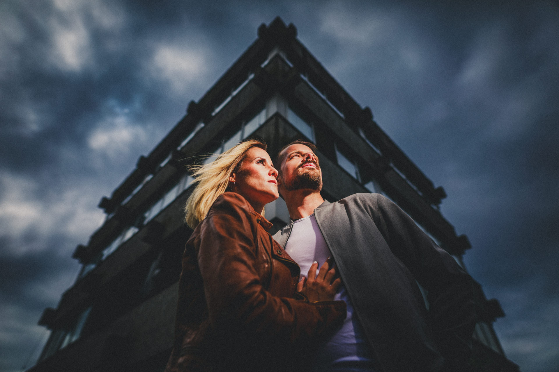 Kristina und Markus stehen eng beieinander bei einem Paarshooting im Lost Place vor einem dunklen Gebäude und schwerem Himmel