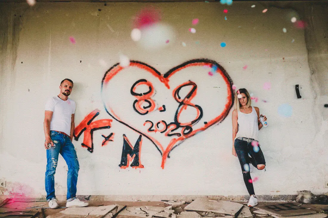 Kristina und Markus stehen bei einem Paarshooting im Lost Place vor einem Graffiti-Herz mit Hochzeitsdatum, während Konfetti im Vordergrund rieselt
