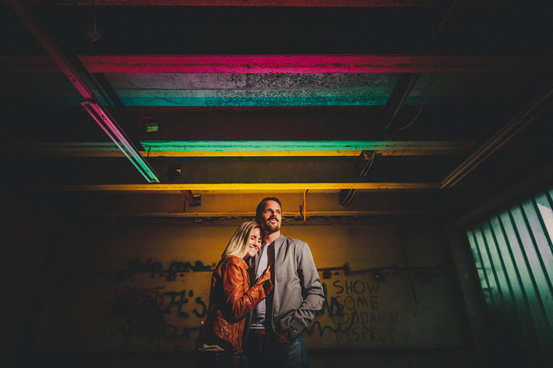 Kristina und Markus stehen unter buntem Blitzlicht in einem dunklen Lost Place mit sichtbaren Deckenbalken und Graffiti