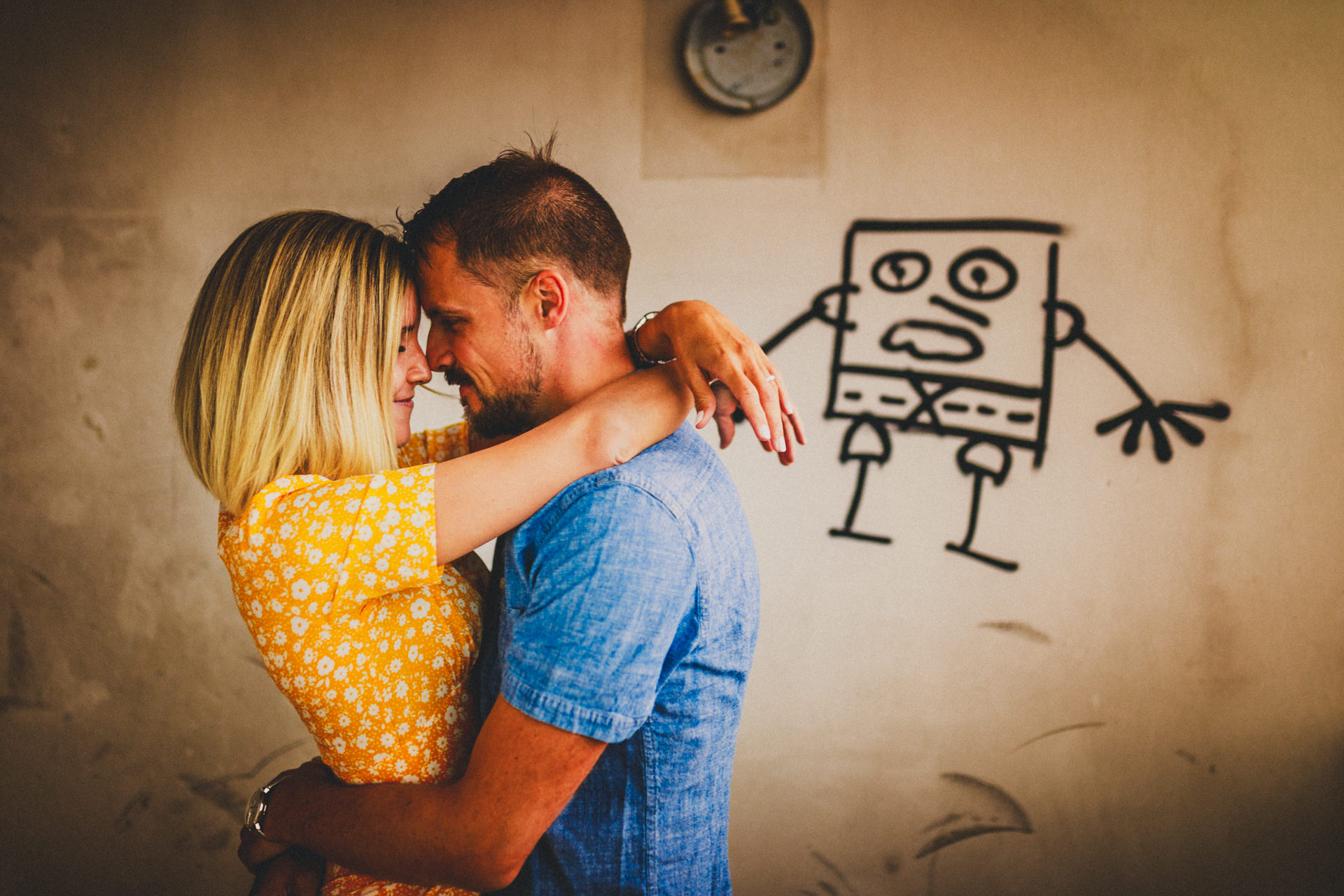 Kristina und Markus umarmen sich eng vor einer Wand mit Graffiti-Figur in einem verlassenen Gebäude