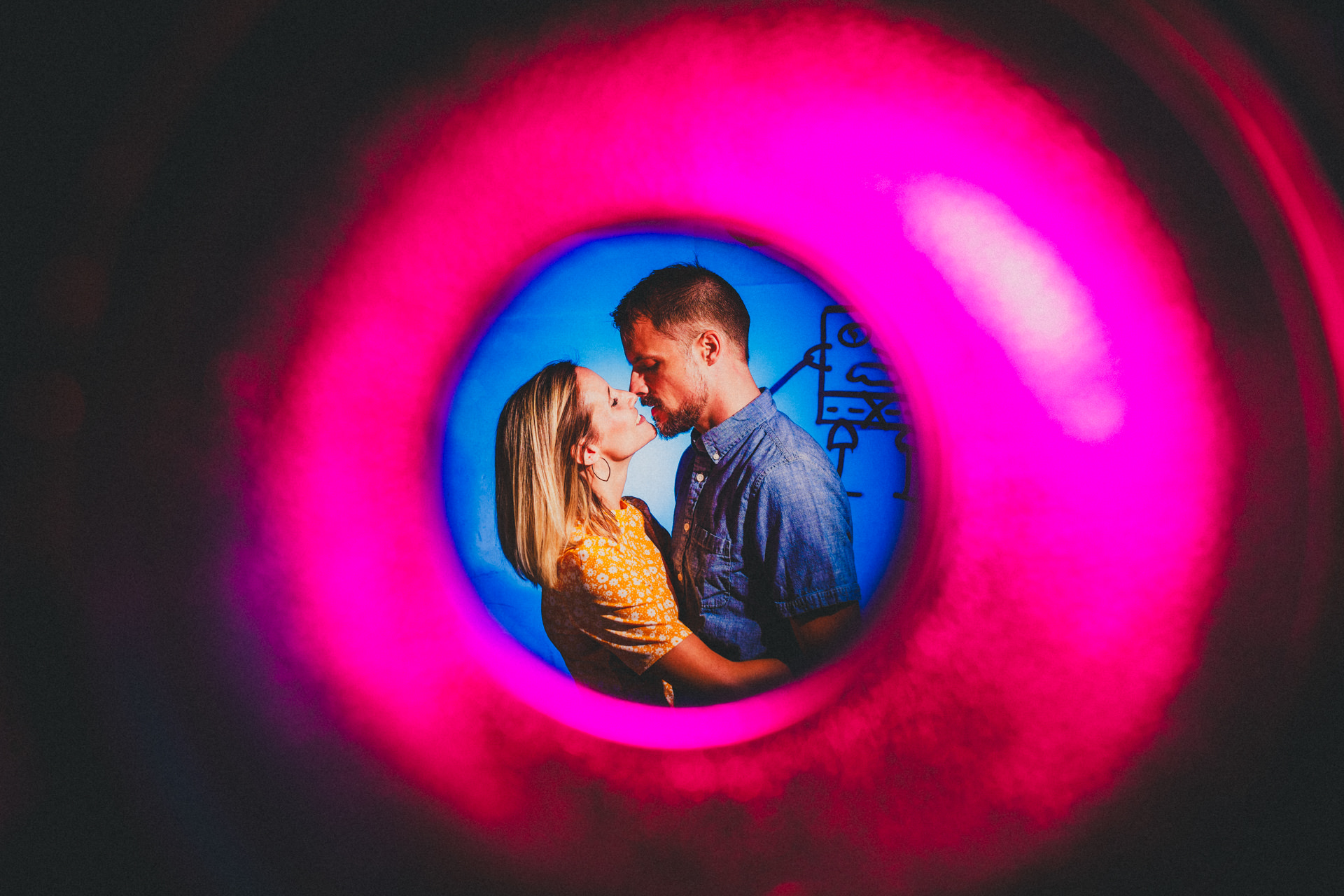 Kristina und Markus stehen sich nah in einem blauen Lichtkreis mit pinkem Neonrahmen in einem Lost Place gegenüber