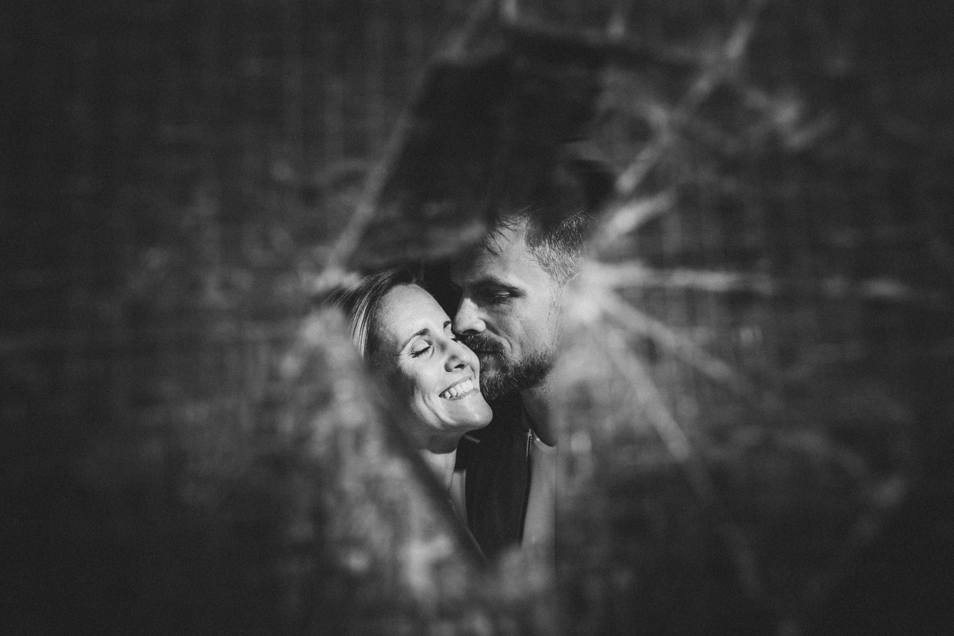 Kristina und Markus lachen eng umschlungen hinter einer beschädigten Oberfläche in Schwarzweiß