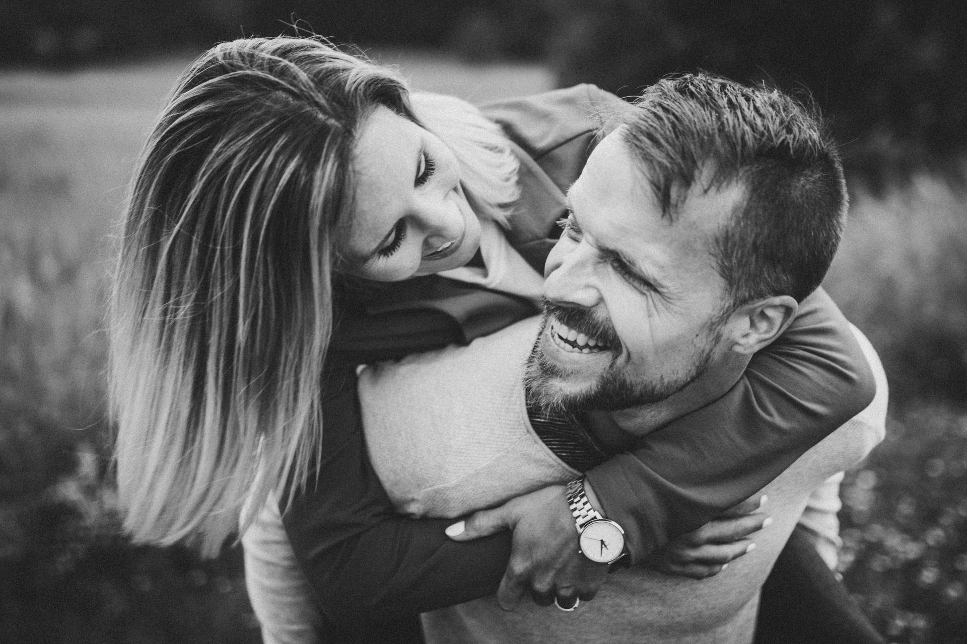 Kristina umarmt Markus lachend von hinten während beide draußen in Schwarzweiß fotografiert werden