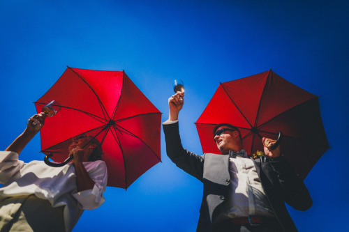 Hochzeitspaar prostet den Gästen mit Gläsern unter roten Schirmen zu