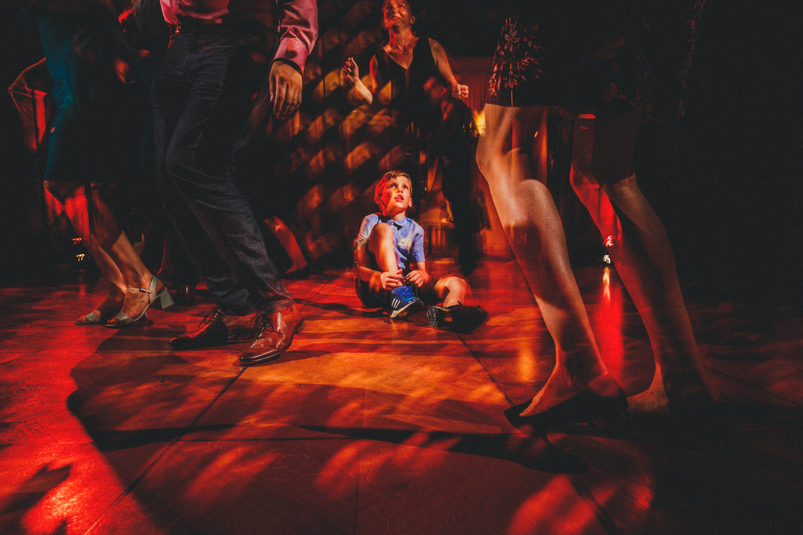 Kind sitzt auf der Tanzfläche zwischen tanzenden Gästen im warmen Licht - fotografiert von Rossi, Hochzeitsfotograf Giessen